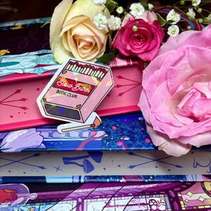 May include: A stack of books with a pink, blue, and purple patterned cover. A holographic sticker with the text "Slow Burn Book Club" and a depiction of matches is on the top book.
