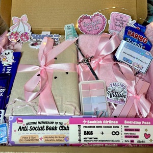 May include: A pink and brown gift box filled with various items, including a book, a pink fluffy notebook, a silver sword, and a sticker that says "Bookish Girlie". The box also includes a pink and white membership card to the "Anti Social Book Club" and a pink and white boarding pass for "Bookish Airlines".