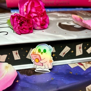 Op de afbeelding: Een holografische sticker van een schattig spook dat een boek leest met de tekst "Bookish Ghouls" op de kaft. De sticker zit op een stapel boeken met roze anjers erop.