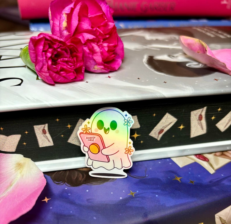 Op de afbeelding: Een holografische sticker van een schattig spook dat een boek leest met de tekst "Bookish Ghouls" op de kaft. De sticker zit op een stapel boeken met roze anjers erop.