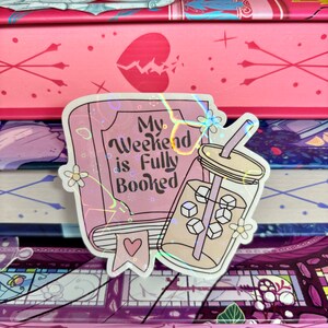 May include: A holographic sticker featuring a pink book and a glass of iced tea. The book reads "My Weekend is Fully Booked".