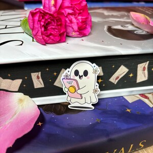Op de afbeelding: Een witte geesten sticker met een roze boek waarop "Bookish Ghoul" staat en een vrolijke uitdrukking. De sticker zit op een stapel boeken met een zwart-wit patroon en een blauw-paarse achtergrond.