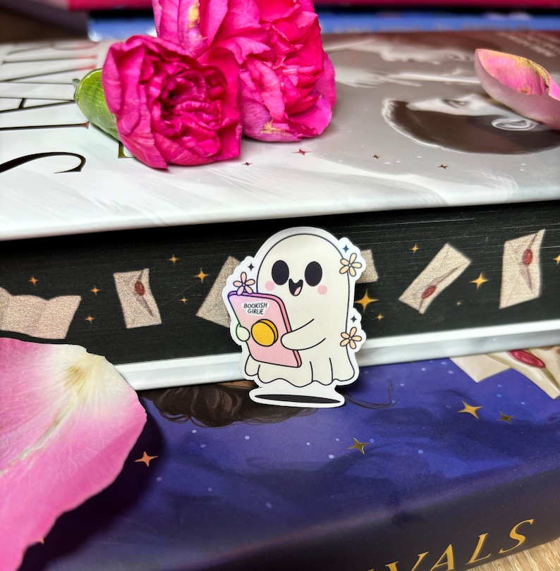 Op de afbeelding: Een witte geesten sticker met een roze boek waarop "Bookish Ghoul" staat en een vrolijke uitdrukking. De sticker zit op een stapel boeken met een zwart-wit patroon en een blauw-paarse achtergrond.