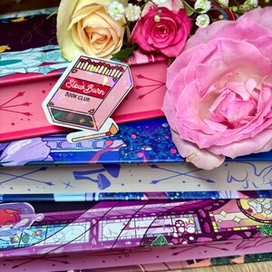 May include: A pink and iridescent sticker with the text "Slow Burn Book Club" and a graphic of a box of matches. The sticker is on top of a stack of books with floral patterned covers.
