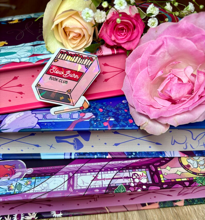 May include: A pink and iridescent sticker with the text "Slow Burn Book Club" and a graphic of a box of matches. The sticker is on top of a stack of books with floral patterned covers.