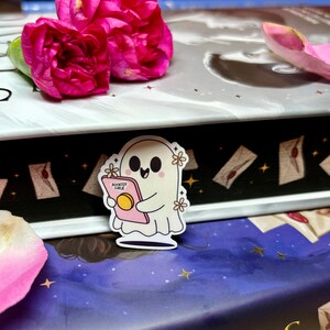 Op de afbeelding: Een witte geesten sticker met een roze boek waarop "Bookish Ghoul" staat en witte bloemen. De sticker zit op een boek met een zwart-wit patroon.