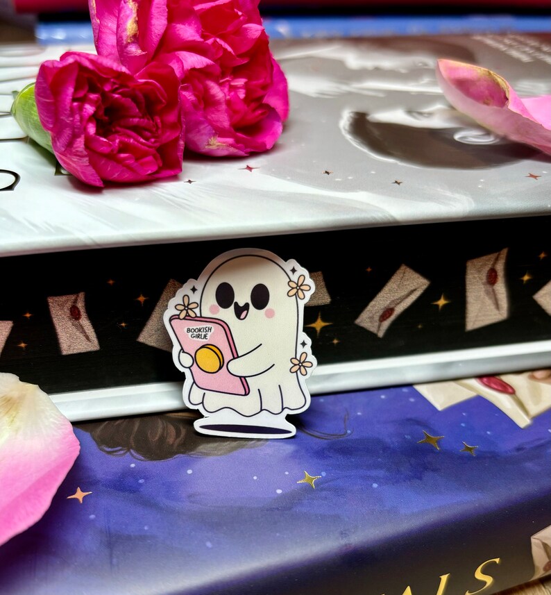Op de afbeelding: Een witte geesten sticker met een roze boek waarop "Bookish Ghoul" staat en witte bloemen. De sticker zit op een boek met een zwart-wit patroon.