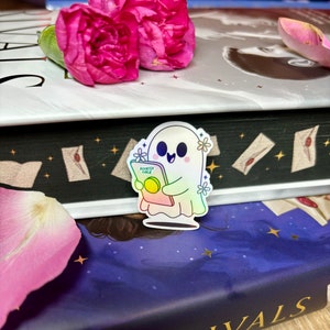Op de afbeelding: Een holografische sticker van een geest die een boek vasthoudt met de tekst "Bookish Ghoul" op de omslag. De sticker zit op een stapel boeken met een roze bloem erop.