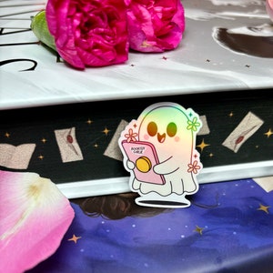 Op de afbeelding: Een holografische sticker van een geest die een boek vasthoudt met de tekst "Bookish Girl" op de omslag. De sticker zit op een boek met een zwarte achtergrond en sterren.