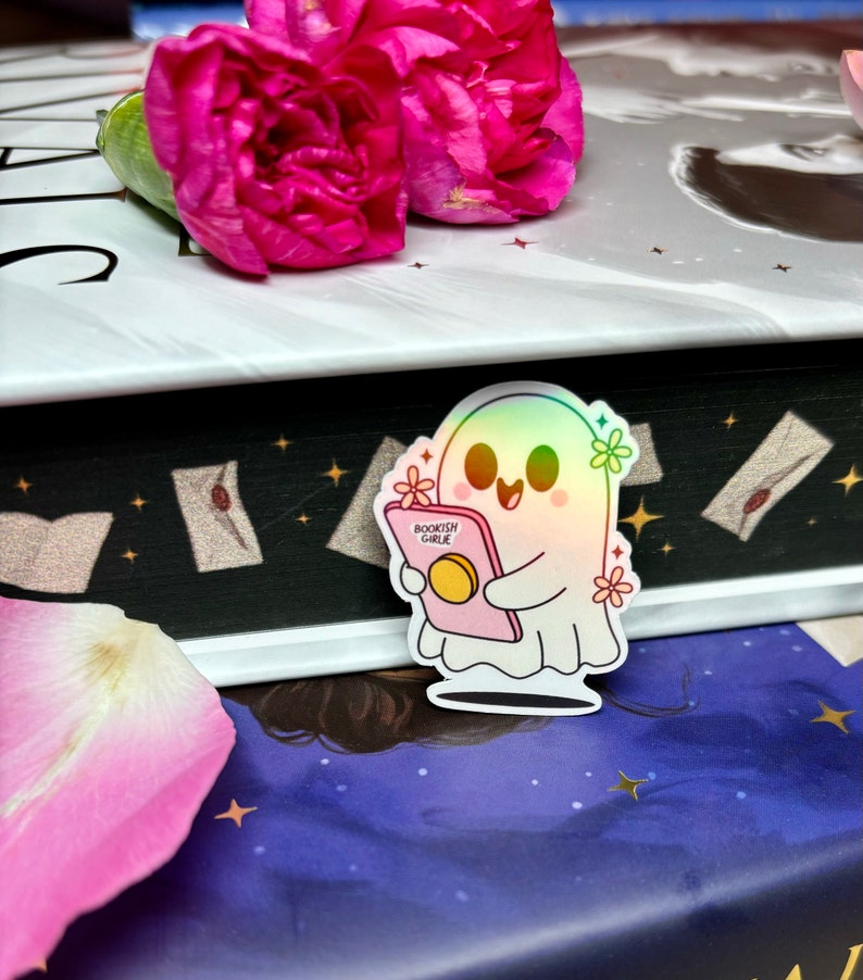 Op de afbeelding: Een holografische sticker van een geest die een boek vasthoudt met de tekst "Bookish Girl" op de omslag. De sticker zit op een boek met een zwarte achtergrond en sterren.