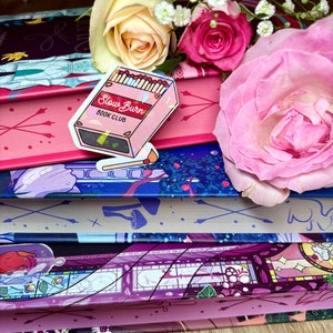 May include: A stack of books with a pink and blue floral design. The top book has a sticker that says "Slow Burn Book Club" with a graphic of matches.