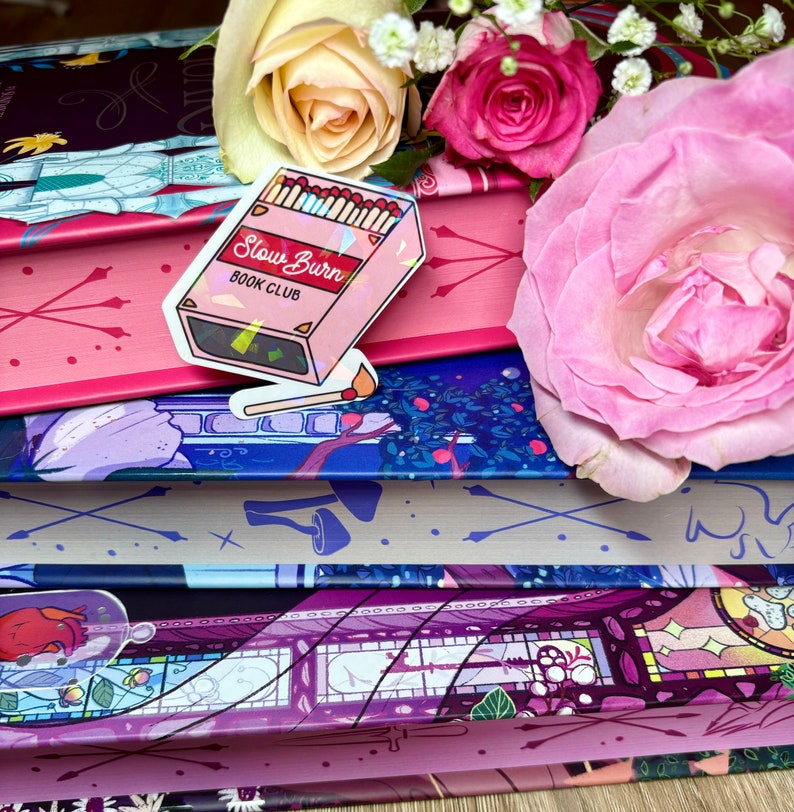 May include: A stack of books with a pink and blue floral design. The top book has a sticker that says "Slow Burn Book Club" with a graphic of matches.