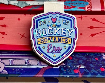 Może przedstawiać: Błyszczący zakładek w kolorze niebieskim i różowym z tekstem "In My Hockey Romance Era" i grafiką hokejowej kija i krążka.