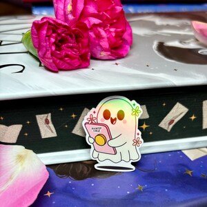 Op de afbeelding: Een holografische sticker van een geest die een roze boek vasthoudt met de tekst "Bookish Girlie" erop. De geest heeft een roze halo en is omringd door roze bloemen.