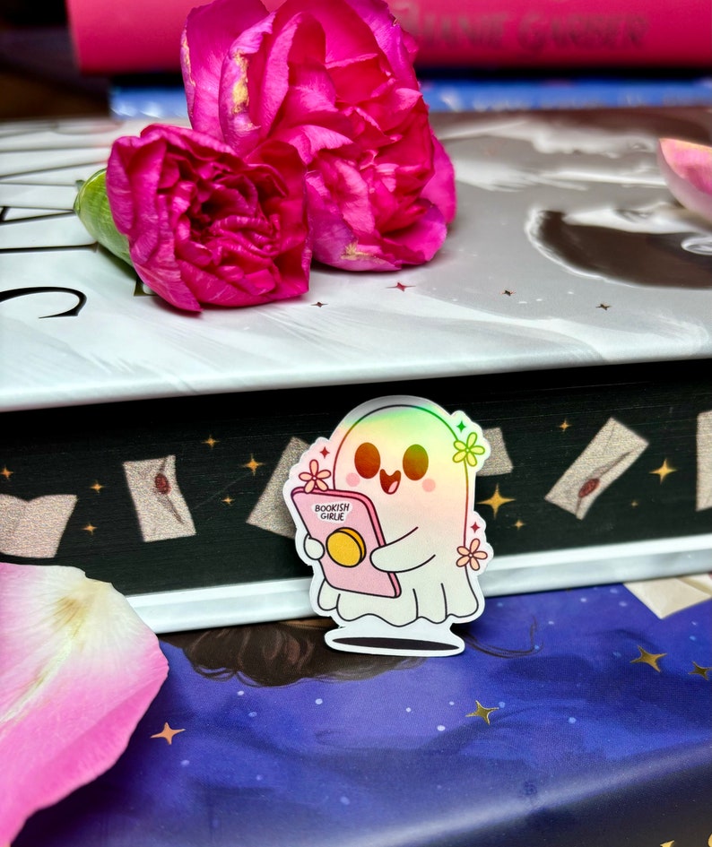 Op de afbeelding: Een holografische sticker van een geest die een roze boek vasthoudt met de tekst "Bookish Girlie" erop. De geest heeft een roze halo en is omringd door roze bloemen.