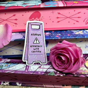 May include: A purple and white caution sign sticker with the text "Reading Approach with Caution" and a warning symbol. The sticker is on top of a stack of books with a pink rose.