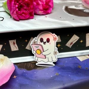 Op de afbeelding: Een witte geesten sticker met een roze boek waarop "BOOKISH GIRLIE" staat en bloemen. De sticker zit op een boek met een donkerblauwe kaft en gouden sterren.