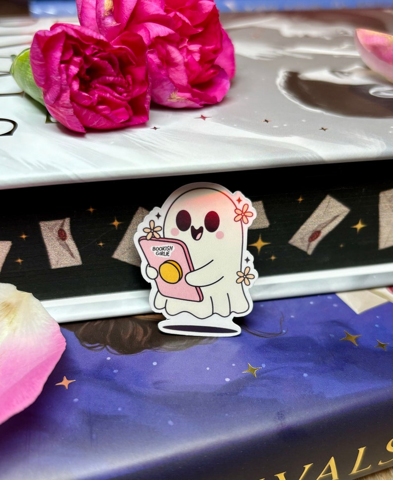 Op de afbeelding: Een witte geesten sticker met een roze boek waarop "BOOKISH GIRLIE" staat en bloemen. De sticker zit op een boek met een donkerblauwe kaft en gouden sterren.