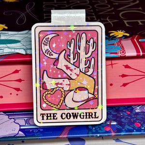 May include: A pink and white bookmark with a glittery finish. The bookmark features a cowgirl wearing a cowboy hat and boots, with a heart and a cactus in the background. The text "THE COWGIRL" is printed at the bottom of the bookmark.