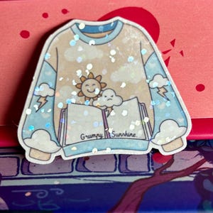 May include: A sticker in the shape of a light yellow sweatshirt with blue sleeves. The design features a smiling sun, a grumpy cloud, and the text "Grumpy Sunshine". The sticker is decorated with glitter.