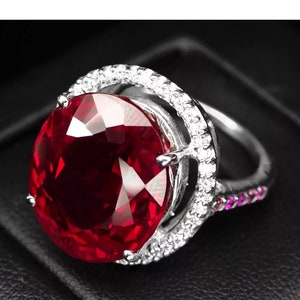 May include: A silver ring featuring a large, oval-shaped ruby gemstone encircled by smaller diamonds. The ring boasts a delicate, intricate design.