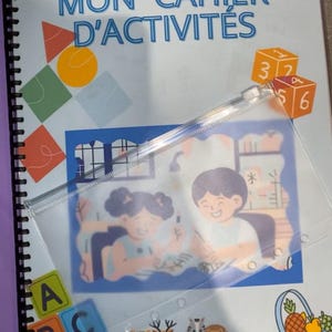 Peut inclure: Un cahier d'activités bleu et blanc avec le titre "MON CAHIER D'ACTIVITES" sur la couverture. La couverture présente également des formes géométriques colorées, une poche en plastique transparente avec une image de deux enfants et un bloc d'alphabet coloré.