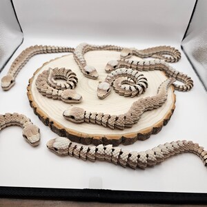 May include: A collection of articulated snake toys in various poses, displayed on a wooden slice. The snakes are primarily beige and brown, with detailed segmented bodies and realistic head designs. Some are coiled, while others are extended.