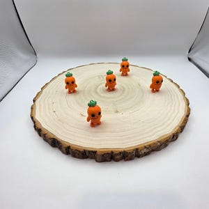 May include: Six small orange carrot-shaped figurines with green tops are arranged on a wooden round slice. The figurines have small black eyes and a simple smile.