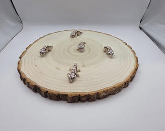 May include: Six small, silver-colored lizard figurines are arranged on a round, light-brown wooden slice with a dark-brown bark edge. The lizards are evenly spaced, creating a circular pattern. The background is white.