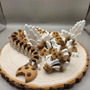 May include: A 3D-printed articulated dragon figurine, featuring a cookie-themed design. The dragon's body is brown with chocolate chip details, and white accents. The dragon is posed on a wooden slice.