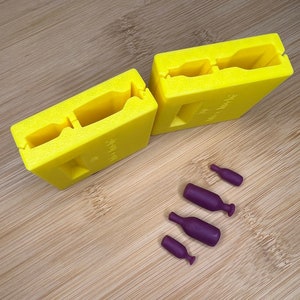 Może przedstawiać: Dwa żółte plastikowe formy do robienia małych butelek, z czterema fioletowymi gumowymi butelkami w różnych rozmiarach.