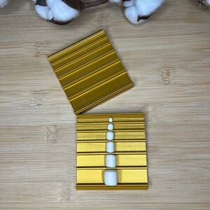 Könnte beinhalten: Ein goldfarbenes Tablett mit sechs Schlitzen zum Aufbewahren von Nagelspitzen. Das Tablett ist geöffnet und ein Stapel von fünf weißen Nagelspitzen befindet sich im mittleren Schlitz.