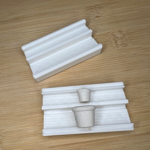 Op de afbeelding: Twee witte keramische potten, waarvan er één op de andere is gestapeld, staan in een witte kunststof mal. De mal heeft een getextureerd oppervlak en is ontworpen om de potten op hun plaats te houden.