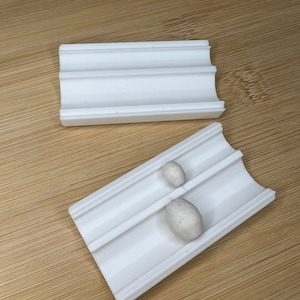 Puede incluir: Dos bandejas de plástico blanco con crestas. La bandeja superior está vacía. La bandeja inferior tiene dos objetos ovalados blancos en las ranuras.