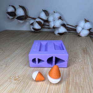 Könnte beinhalten: Lilafarbene Silikonform mit zwei rechteckigen und drei kegelförmigen Vertiefungen. Zwei orangefarbene und weiße kegelförmige Polymer Clay-Stücke liegen neben der Form.