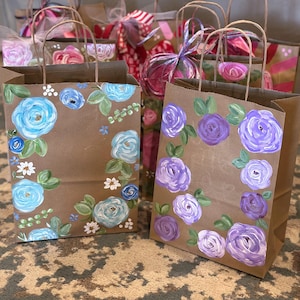 Pode incluir: Sacos de presente de papel castanho com desenhos florais pintados à mão. Uma bolsa apresenta rosas azuis claras, enquanto outra mostra rosas roxas. Ambas as bolsas têm folhas verdes e detalhes brancos, com alças de papel castanho.