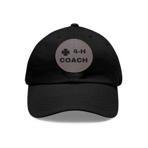 May include: A black baseball cap featuring a circular grey patch centered on the front. The patch displays a black four-leaf clover icon followed by the text 4-H COACH in bold, dark lettering. The cap has a curved brim and stitched detailing.