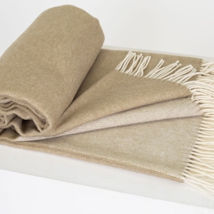 Op de afbeelding: Een zachte, beige deken met een subtiel ombre-effect, voorzien van witte franjes aan alle kanten. De deken is opgerold en gepresenteerd op een wit oppervlak.