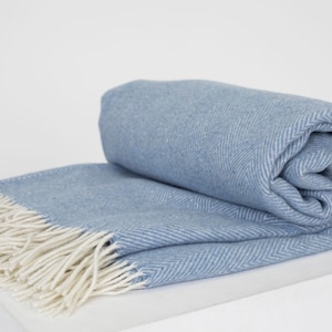 Könnte beinhalten: Eine weiche, blaue Wolldecke mit weißem Fransenbesatz. Die Decke hat ein Fischgrätenmuster und ist in der Hälfte gefaltet.