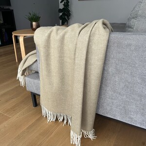 May include: A beige throw blanket with white fringe draped over a gray couch. The blanket is made of a soft, textured fabric.