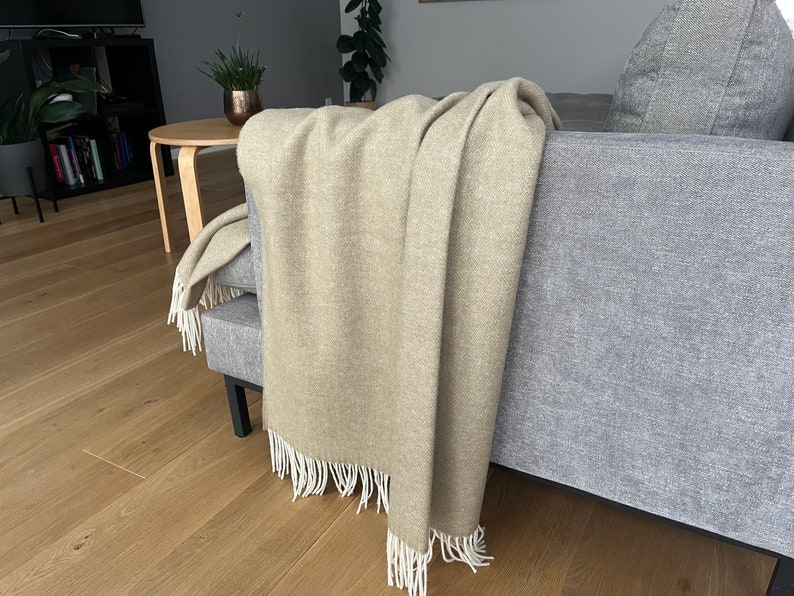 May include: A beige throw blanket with white fringe draped over a gray couch. The blanket is made of a soft, textured fabric.