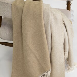 May include: A beige and cream throw blanket with fringe edges draped over a light beige armchair.
