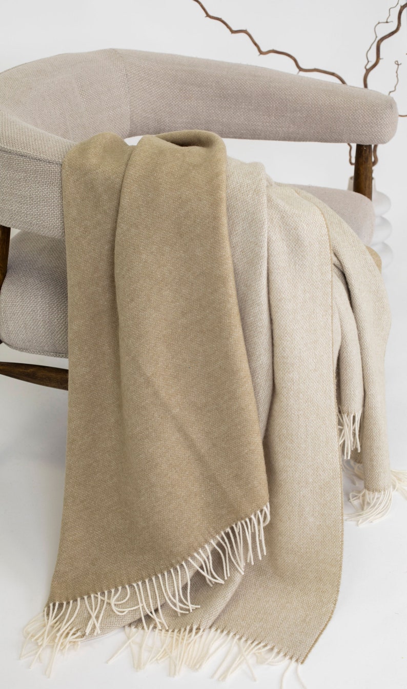 May include: A beige and cream throw blanket with fringe edges draped over a light beige armchair.