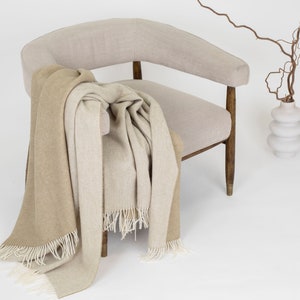 May include: A beige armchair with a brown and beige throw blanket draped over it. A white vase with a brown branch is in the background.