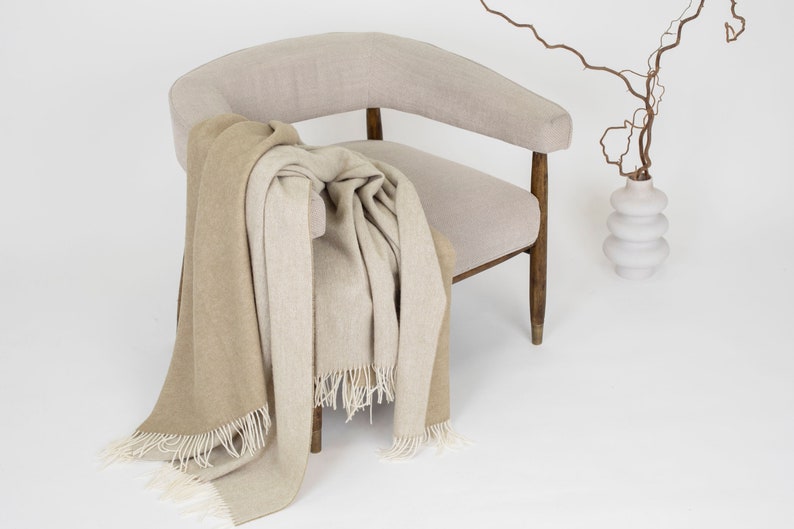 May include: A beige armchair with a brown and beige throw blanket draped over it. A white vase with a brown branch is in the background.