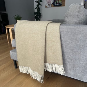 May include: A beige throw blanket with white fringe is draped over a gray couch. The blanket is folded in half and the fringe is visible on both sides.