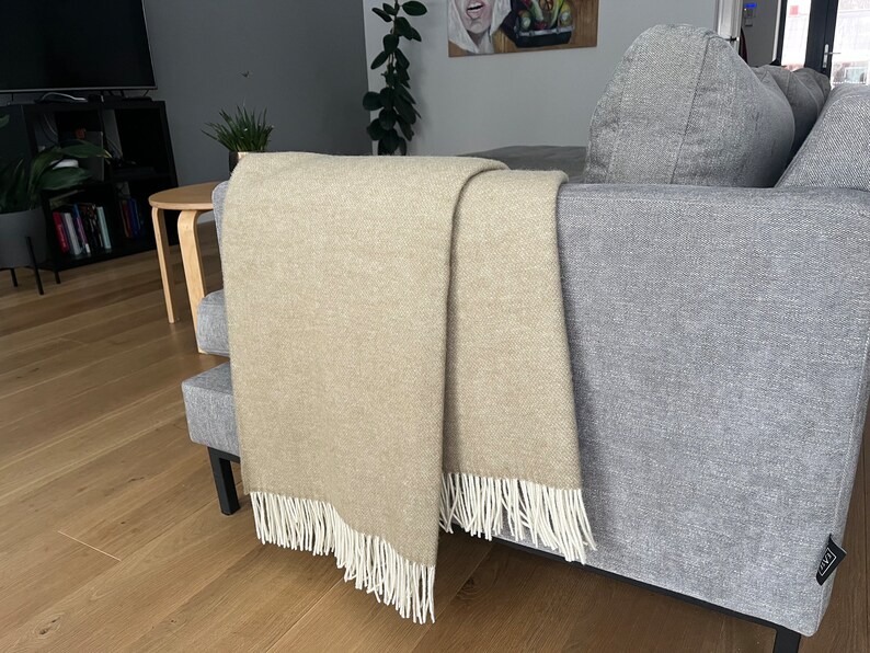 May include: A beige throw blanket with white fringe is draped over a gray couch. The blanket is folded in half and the fringe is visible on both sides.