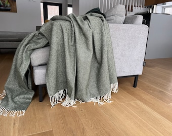 May include: A sage green throw blanket featuring a woven herringbone pattern and white fringe trim along the edges. The blanket is draped over a light gray sofa, showcasing the textured fabric and the detailed weave of the material.