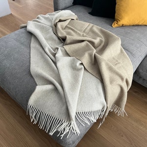 May include: A soft, two-toned throw blanket with a light beige and gray color scheme. The blanket has a fringe border and is draped over a gray couch.