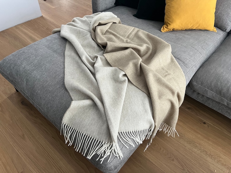 May include: A soft, two-toned throw blanket with a light beige and gray color scheme. The blanket has a fringe border and is draped over a gray couch.